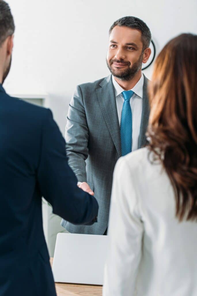 selective focus of handsome advisor in suit shaking hands with investors at office